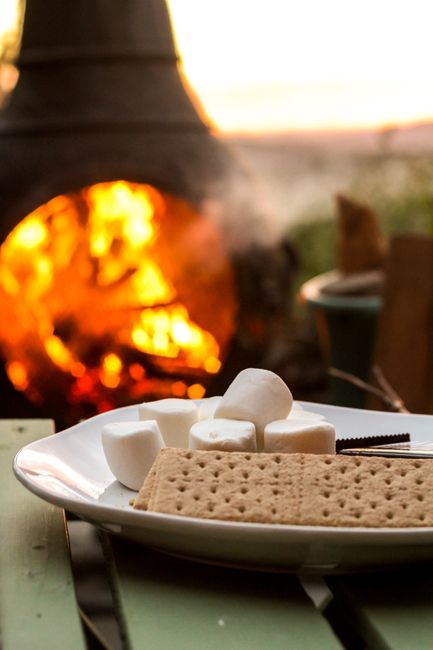 A white ceramic plate holds several large, rounded marshmallows and a rectangular, plain biscuit cracker, arranged on a table outdoors during sunset. In the background, a brick or stone outdoor oven or fireplace with a glowing fire is visible, emitting warm light and smoke, suggesting a cozy, outdoor setting. To the right, there is a small table with a plant in a pot. The scene captures a casual moment likely related to a home or outdoor gathering, with a focus on the snack presentation and the ambient glow of the fire, aligned with activities such as unpacking or preparing for a meal during a household move or outdoor event, relevant to home relocation and outdoor furniture transport services provided by Man with Van Grahame Park.