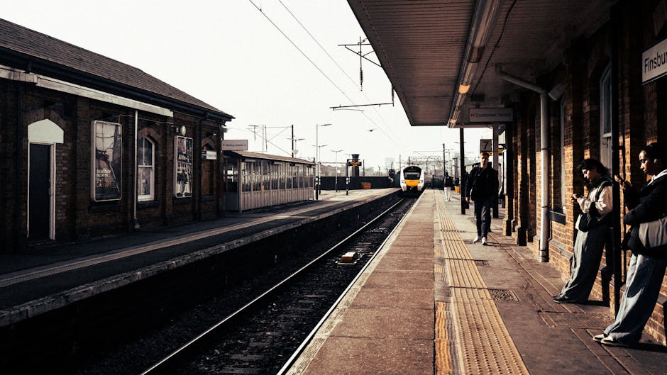 A train station platform with a yellow and white train approaching in the distance. The platform has a tiled surface with tactile paving near the edge for visually impaired passengers. Several people are standing along the platform, some near the brick station building on the right, which features a sign indicating a train stop. Inside the station building, visible through windows, are posters and advertisements. Overhead, there are electrical lines and signals associated with the railway. The scene is well-lit with natural daylight, illustrating an active setting for home relocation or furniture transport activities, where careful loading and waiting processes are part of the moving logistics supported by Man with Van Grahame Park.