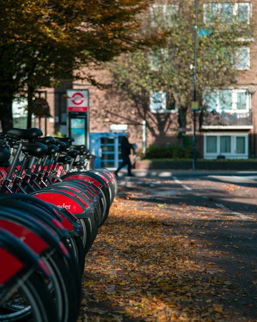A row of red and black bicycles parked in a designated bike-sharing station along a paved sidewalk covered with fallen autumn leaves, with a blue public telephone kiosk, a bench, and a modern brick apartment building with large windows in the background. In the distance, a person dressed in dark clothing is walking near a tall tree with multicoloured foliage, casting shadows on the pavement. The scene is captured during daylight hours, with natural light illuminating the surroundings. The image depicts an urban residential environment, relevant to house removals and moving logistics services offered by Man with Van Grahame Park, highlighting the importance of parking and access considerations near Colindale Station, GRAHAME PARK for effective furniture transport and home relocation activities.