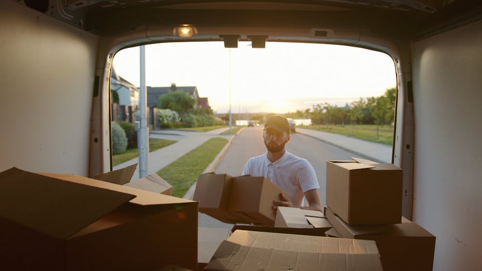 A man wearing a white polo shirt and glasses is seen loading cardboard boxes into the back of a moving van during a home relocation. The van's interior is visible with several packed cardboard boxes o