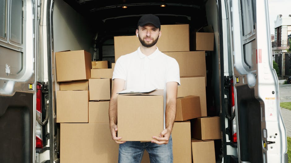 A man wearing a white polo shirt and glasses is seen loading cardboard boxes into the back of a moving van during a home relocation. The van's interior is visible with several packed cardboard boxes o