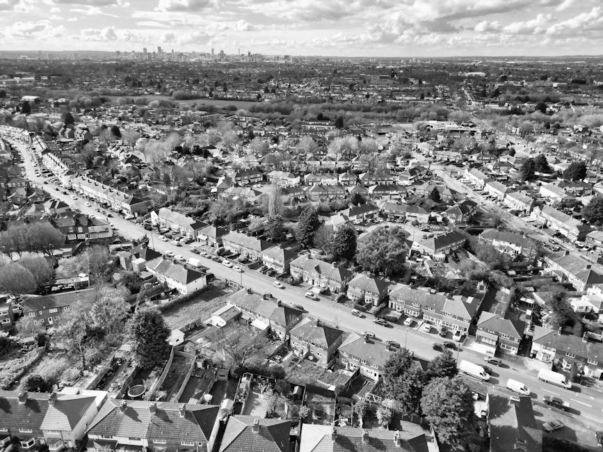 Aerial black-and-white photograph of a residential neighbourhood in Grahame Park, NW9, showing rows of houses with pitched roofs, enclosed gardens, and tree-lined streets. Several cars are parked along the curbs, with one vehicle visible on the road indicating transportation activity. The scene includes backyards with fencing, and some houses have visible driveways or yards filled with various items. In the distance, the skyline of London is faintly visible under a partly cloudy sky. This image captures the typical setting for house removals and furniture transport activity, illustrating the environment where relocation and packing processes by companies like Man with Van Grahame Park may take place.