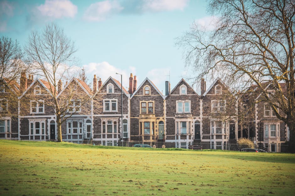 A row of several Victorian-style terraced houses made of grey brick, with large bay windows and pitched roofs, situated along a residential street in Grahame Park. The houses are positioned behind a fenced boundary, with leafless trees lining the front of the properties, indicating a colder season. The foreground features a green, grassy area with a few scattered leaves, and the sky above is partly cloudy with patches of blue. Visible in the image are various cardboard boxes, furniture blankets, and packing materials placed near the entrances, suggesting preparations for home relocation. A man with a blue jacket is seen loading or unloading a large cardboard box from a moving van parked on the pavement, with additional boxes and household items visible inside the van, which is partially out of view. This scene illustrates the physical process of furniture transport and packing during residential removals, relevant to services offered by Man with Van Grahame Park as part of a home moving or packing and moving logistics.