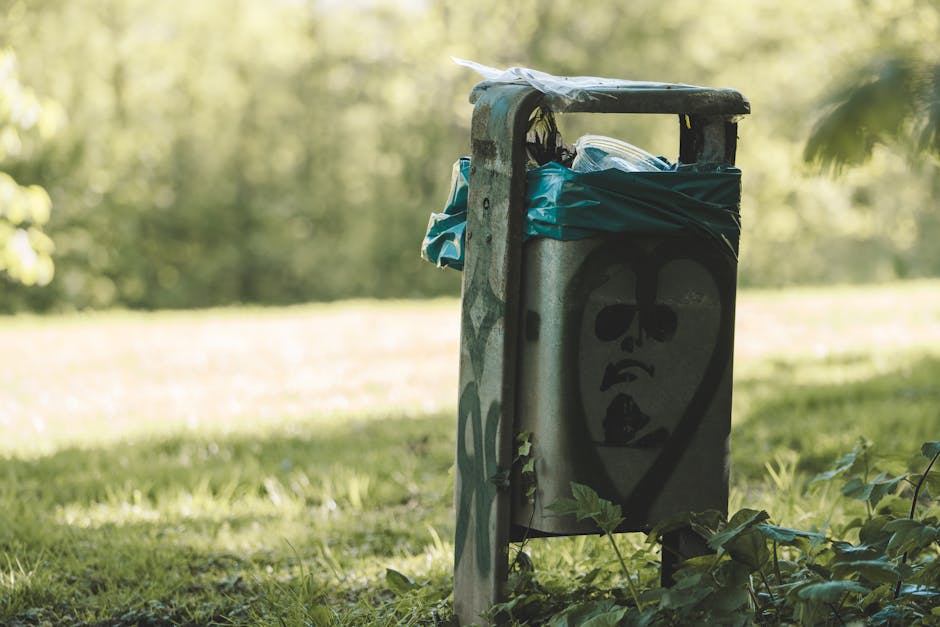 A weathered public waste collection bin standing on a grassy area outdoors, attached to a metal post with visible graffiti artwork depicting a stylized face with sunglasses and an open mouth. The bin has a black, worn lid, and is lined with a blue plastic bag overflowing with miscellaneous trash, including cardboard and plastic packaging. The background features blurred green foliage and sunlight filtering through trees, indicating a park or open green space. The scene suggests the disposal of waste, relevant to house removals and home relocation services, with the bin positioned near a pathway or outdoor area where items may be grouped for collection or transportation, typical of loading and moving logistics as seen in professional removals by [COMPANY_NAME].