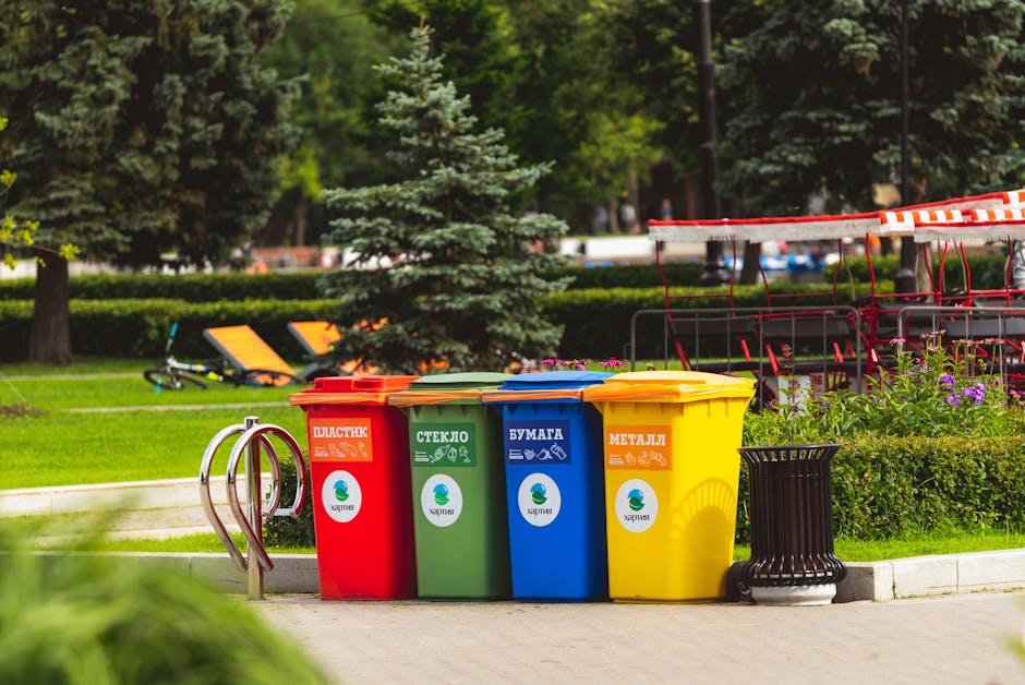 A weathered public waste collection bin standing on a grassy area outdoors, attached to a metal post with visible graffiti artwork depicting a stylized face with sunglasses and an open mouth. The bin has a black, worn lid, and is lined with a blue plastic bag overflowing with miscellaneous trash, including cardboard and plastic packaging. The background features blurred green foliage and sunlight filtering through trees, indicating a park or open green space. The scene suggests the disposal of waste, relevant to house removals and home relocation services, with the bin positioned near a pathway or outdoor area where items may be grouped for collection or transportation, typical of loading and moving logistics as seen in professional removals by [COMPANY_NAME].