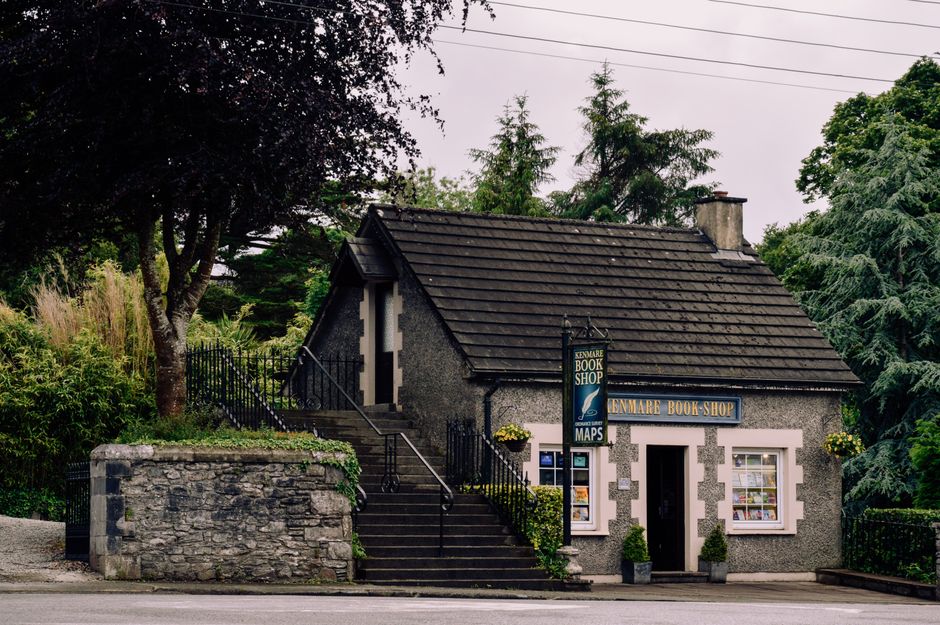 A small, grey stone building with a dark tiled pitched roof situated on a quiet street, surrounded by tall greenery including trees and bushes. The building houses a bookshop, as indicated by the sign hanging from the front wall that reads 'Lenmare Book Shop.' There are two small windows with white frames and flower pots with yellow flowers hanging on either side of the entrance. A black metal fence runs along the side of the property, with a staircase leading from the street level up to the side door, flanked by a stone wall and a set of steps equipped with a handrail. The scene is illuminated by natural daylight, emphasizing the building's exterior and the lush foliage around it. This image captures the environment where house removals or furniture transport services by [COMPANY_NAME], such as loading, loading process, or packing and moving, might take place during a home relocation in Grahame Park.