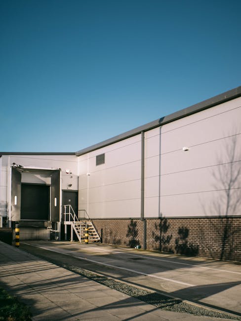 A clear view of the exterior of a large commercial warehouse or distribution centre during daytime, with a wide loading bay featuring an industrial roller shutter door, a small set of metal stairs with handrails leading up to a door, and a paved parking area with marked parking spaces in front. The building has an external wall made of horizontal grey panels and a lower section of brickwork, with shadows of nearby trees cast onto the wall. The bright blue sky overhead indicates good weather, and the scene is illuminated by natural daylight. This setting is typical of a site used for furniture transport and home relocation services, where bulky items are loaded or unloaded into vans. The image aligns with professional removals operations, such as those conducted by Man with Van Grahame Park, supporting the logistics of packing, loading, and transporting belongings during house removals or business relocations.