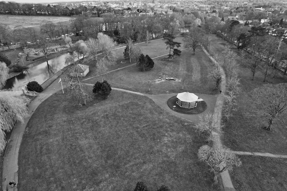 Aerial black-and-white photograph of a park with a large open grassy area, numerous leafless trees, and pathways intersecting the space. A small pond is visible on the left side, surrounded by a walking path and some benches, with a few parked cars nearby. A circular gazebo with a conical roof is situated centrally within a paved area, and the park is bordered by residential buildings and additional greenery in the background. The scene appears quiet with no people present. This image captures the outdoor environment surrounding a community park, illustrating an open space suitable for light outdoor activities, and reflects the tranquil setting for residential relocations in the Grahame Park area. The visual emphasizes the spaciousness and natural features of the park, relevant to types of environments managed by companies like Man with Van Grahame Park during house removals or home relocation projects.