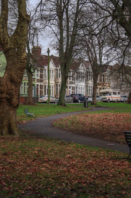 An outdoor scene in a residential area during late autumn with several large, mature trees with bare branches and textured bark lining a paved pathway. Fallen leaves cover parts of the grass and pathway, with some scattered on the benches positioned alongside the walkway. Multiple parked cars are visible along the street behind the trees, including hatchbacks, sedans, and a van, in various colours such as silver, white, and dark shades. In the background, there are traditional three-storey Victorian-style terraced houses with bay windows, brick facades, and pitched roofs, some with chimneys. The overcast sky casts diffuse natural light over the scene. The image captures a quiet, suburban street environment suitable for house removals and property moving services, with the setting indicating a neighbourhood appropriate for residential relocations. Man with Van Grahame Park would use such context to support home relocation logistics and loading processes.