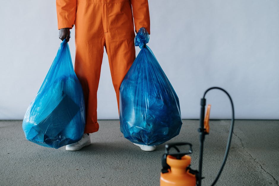 A person dressed in orange work overalls is standing indoors on a carpeted floor, holding two large blue plastic garbage bags filled with items during a home relocation process. The individual's face is not visible, but their hands grip the bags by the tied handles. In front of them, a small orange and black sprayer or cleaning device is positioned on the floor, and behind, there is an open doorway leading to an adjacent room. The scene is set against a plain light grey wall, emphasizing the practical aspects of packing and loading for a removals service. This image reflects the packing stage involved in furniture transport and house removals, consistent with professional moving and waste disposal rules in Barnet, as highlighted on the Man with Van Grahame Park website.
