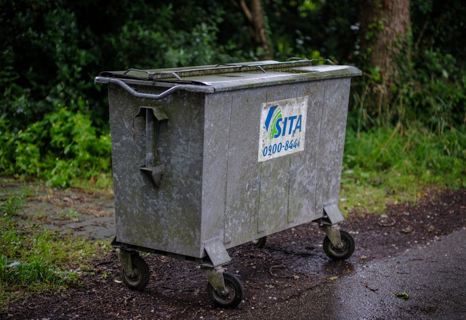A grey, wheeled waste collection container with a partially open lid, positioned on an outdoor pavement surface near a grassy area with green shrubbery and trees in the background. The container has a blue and green SITA logo and a contact number on its side. It is used for general waste disposal during home relocation or moving processes. The container appears weathered and slightly mossy, indicating it has been outdoors for some time, and is situated close to the edge of the property's frontage, ready for collection or transportation as part of an organised removal service by Man with Van Grahame Park. The scene suggests preparation for waste clearance in a move or clearance project, with the container standing on a paved pathway and surrounded by natural greenery, appropriate for a description of loading and waste disposal activities related to house removals and relocation logistics.