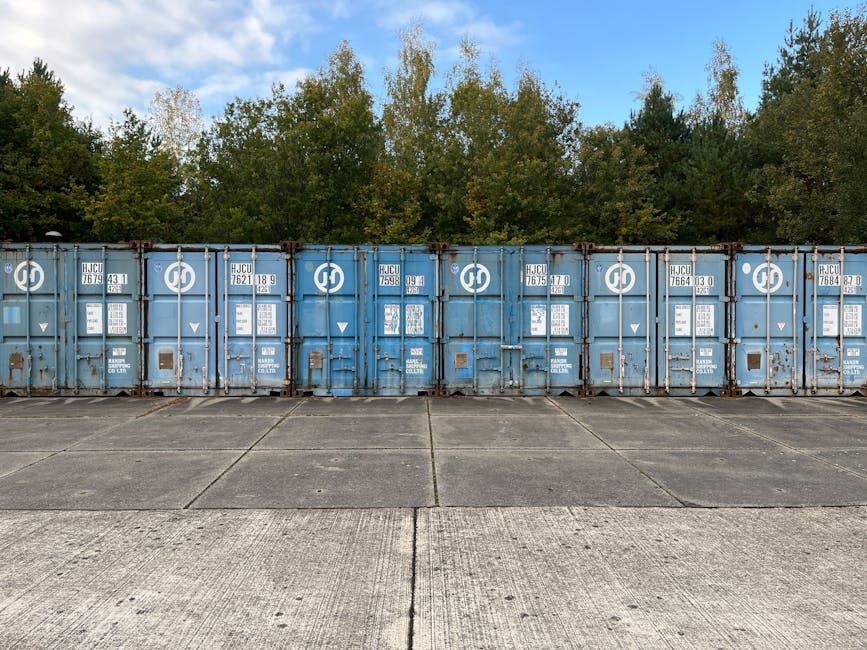 A row of blue shipping containers with visible identification plates, hinges, and locking mechanisms, positioned side by side on a concrete ground surface. The containers are outdoors, with a backdrop of green trees and a partly cloudy sky. The setting suggests a storage or transportation environment, relevant to house removals and moving logistics. In the context of furniture transport or home relocation, the containers may be used for secure storage of household items during the moving process. The image is taken in daylight, highlighting the metal surfaces and the outdoor space, illustrating the staging area for loading or unloading furniture and boxes. This scene aligns with professional removals services provided by Man with Van Grahame Park, emphasizing organized storage and transport solutions for clients near Grahame Park estate (NW9).