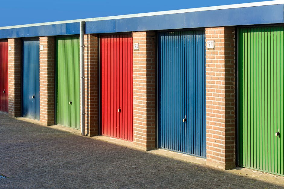 A row of six garage doors situated along a brick building facade with a blue overhang roof, featuring a sequence of red, green, and blue metal doors, each with a small round lock in the center. The doors are positioned on a paved pathway, with some doors having visible small labels and numbers above them. The garage doors are part of a storage facility or garage units, likely used for home relocation or furniture transport. The scene is illuminated by natural daylight, casting shadows on the pavement, and the environment appears clean and orderly. The colorful doors contrast against the brick walls, and a vertical metal pipe runs along the wall between two of the garages. This setting is associated with moving services such as packing, loading, and transportation provided by companies like Man with Van Grahame Park, specializing in house removals and storage solutions.